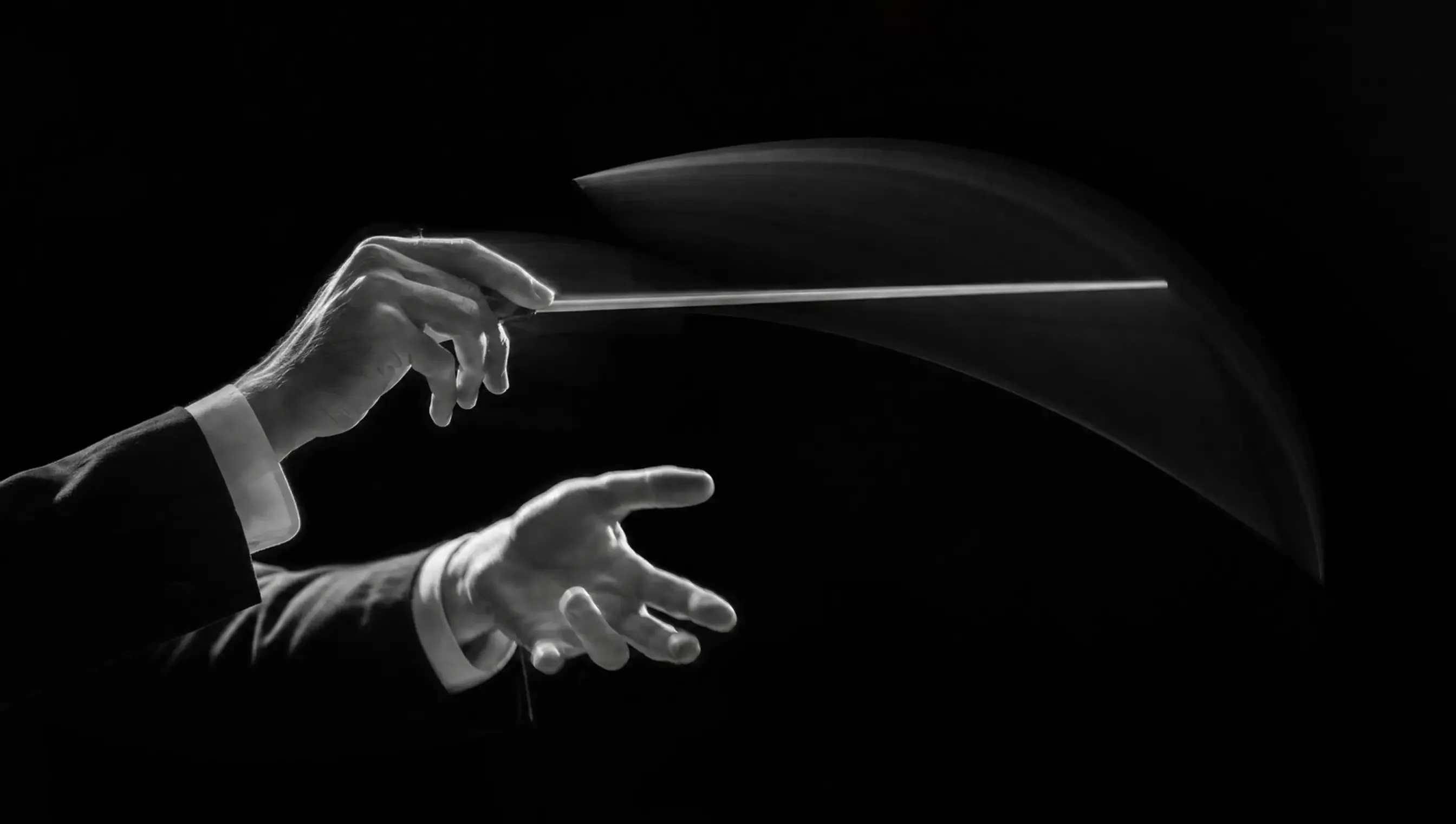 Black and white photograph of a conductor's hands mid-gesture, baton tracing an arc of motion blur.